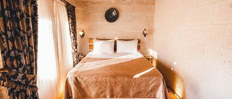 A room with a bed, wooden flooring, stone walls, and a window with curtains.