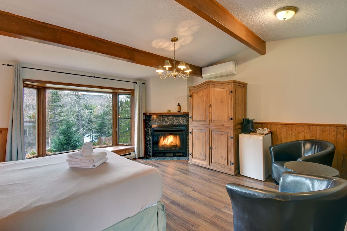 A room with a bed, a fireplace, wooden cabinets, and a view of trees through large windows.