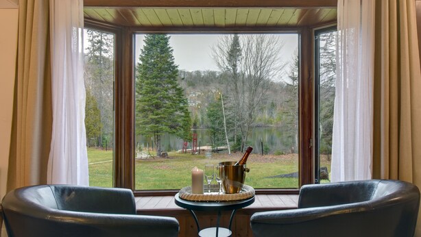 A room with a large window, two chairs, a small table with a candle, glasses, and a champagne bucket. - Hôtel du Lac Lenore (Sainte-Adele)