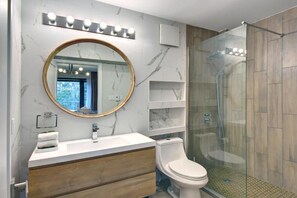 A modern bathroom with a marble vanity, a round mirror, a toilet, and a glass-enclosed shower. - Hôtel du Lac Lenore (Sainte-Adele)