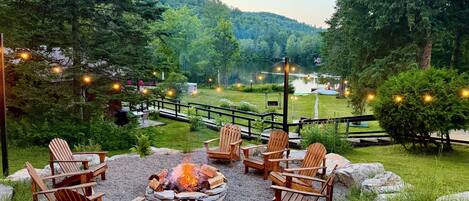 Un espacio al aire libre con una fogata, rodeado de sillas Adirondack, con vistas a un lago y colinas boscosas.