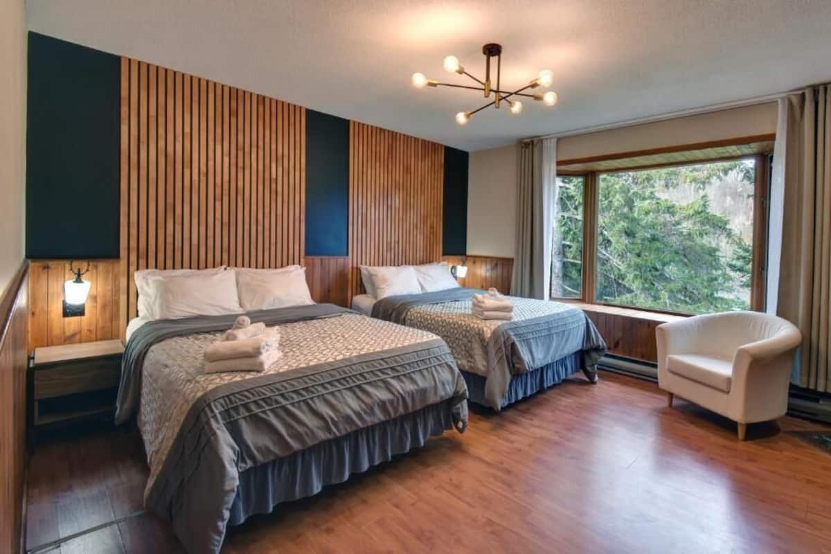 A hotel room with two beds, wooden paneling on the walls, a large window with a view of trees, and a modern chandelier.