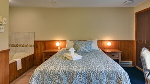 A hotel room with a large bed, wooden headboard, two bedside tables, a nightstand, a chair, and a bathroom with a bathtub and toilet. - Hôtel du Lac Lenore (Sainte-Adele)