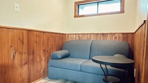 A corner room with a blue sofa, a small round table, wooden paneling, and a window. - Hôtel du Lac Lenore (Sainte-Adele)