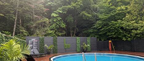 A swimming pool with a wooden deck, surrounded by a fence with greenery.