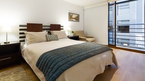 A modern bedroom with a large bed, a nightstand, a bedside table, a chair, and a window with a view of buildings. - Suites Metropoli Edificio Torino (Quito)