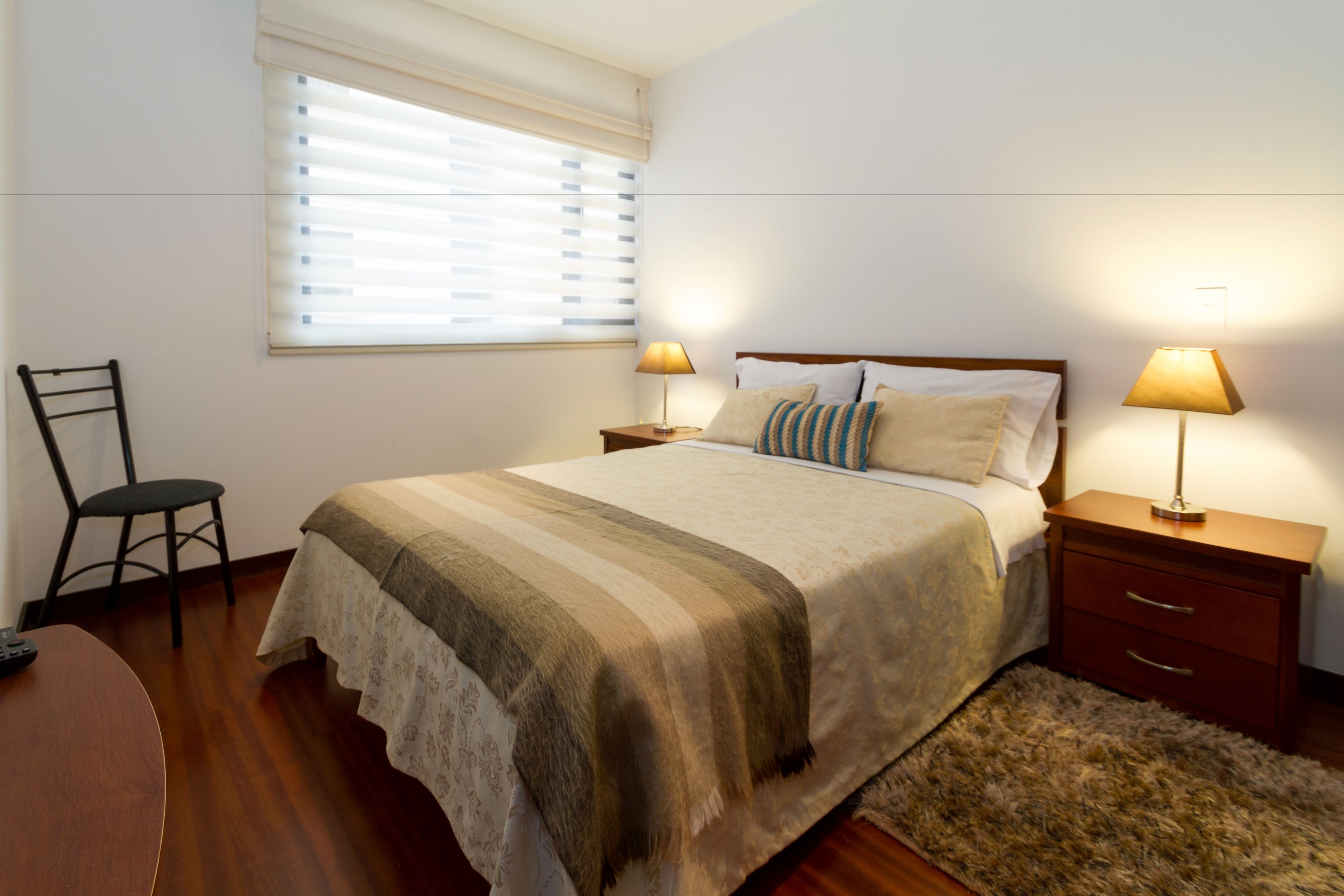 A bedroom with a bed, two bedside tables with lamps, a chair, and a window with blinds.