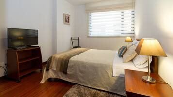 Habitación de hotel con cama, televisión, escritorio con lámpara y ventana con persianas.