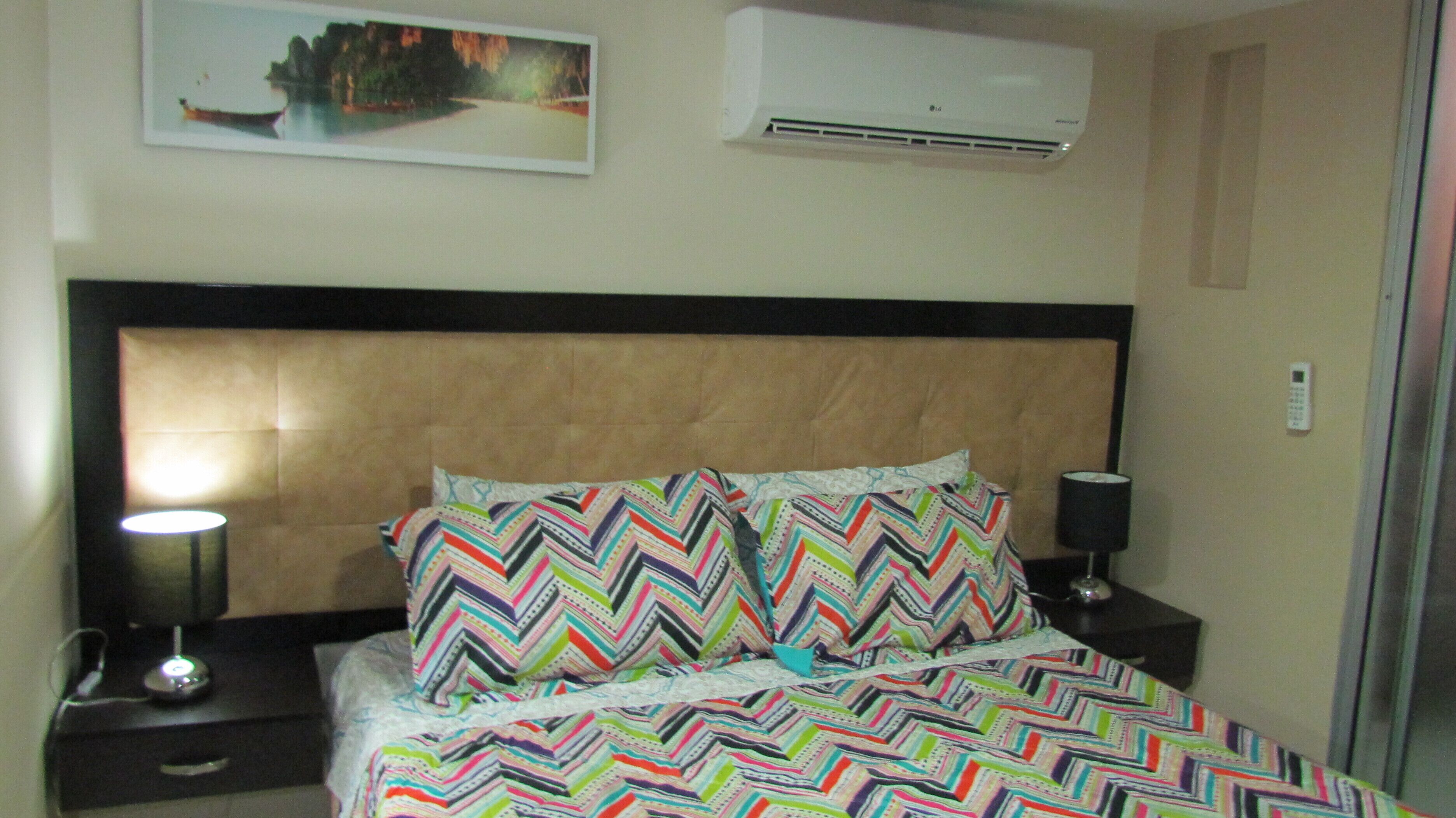 A hotel room with a bed, two bedside tables, a lamp, an air conditioner, and a painting on the wall.