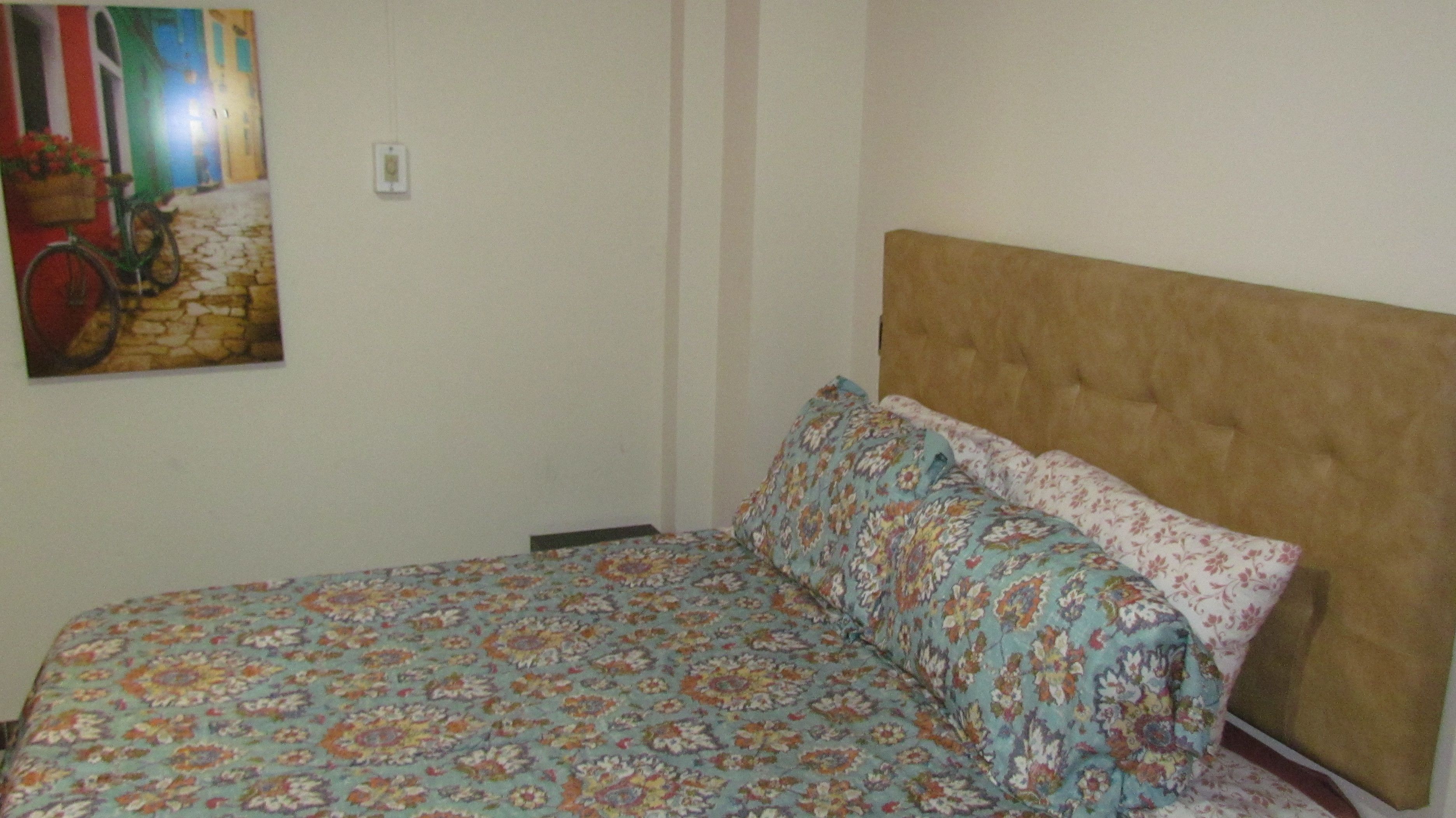 Un dormitorio con colcha estampada, cabecera y un cuadro colgado en la pared que muestra una escena callejera.