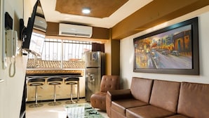 Sala de estar com sofá de couro marrom, mesa de centro e um quadro na parede. Há uma área de cozinha com geladeira, micro-ondas e cafeteira. O quarto possui piso de azulejo e uma janela com vista para o exterior.