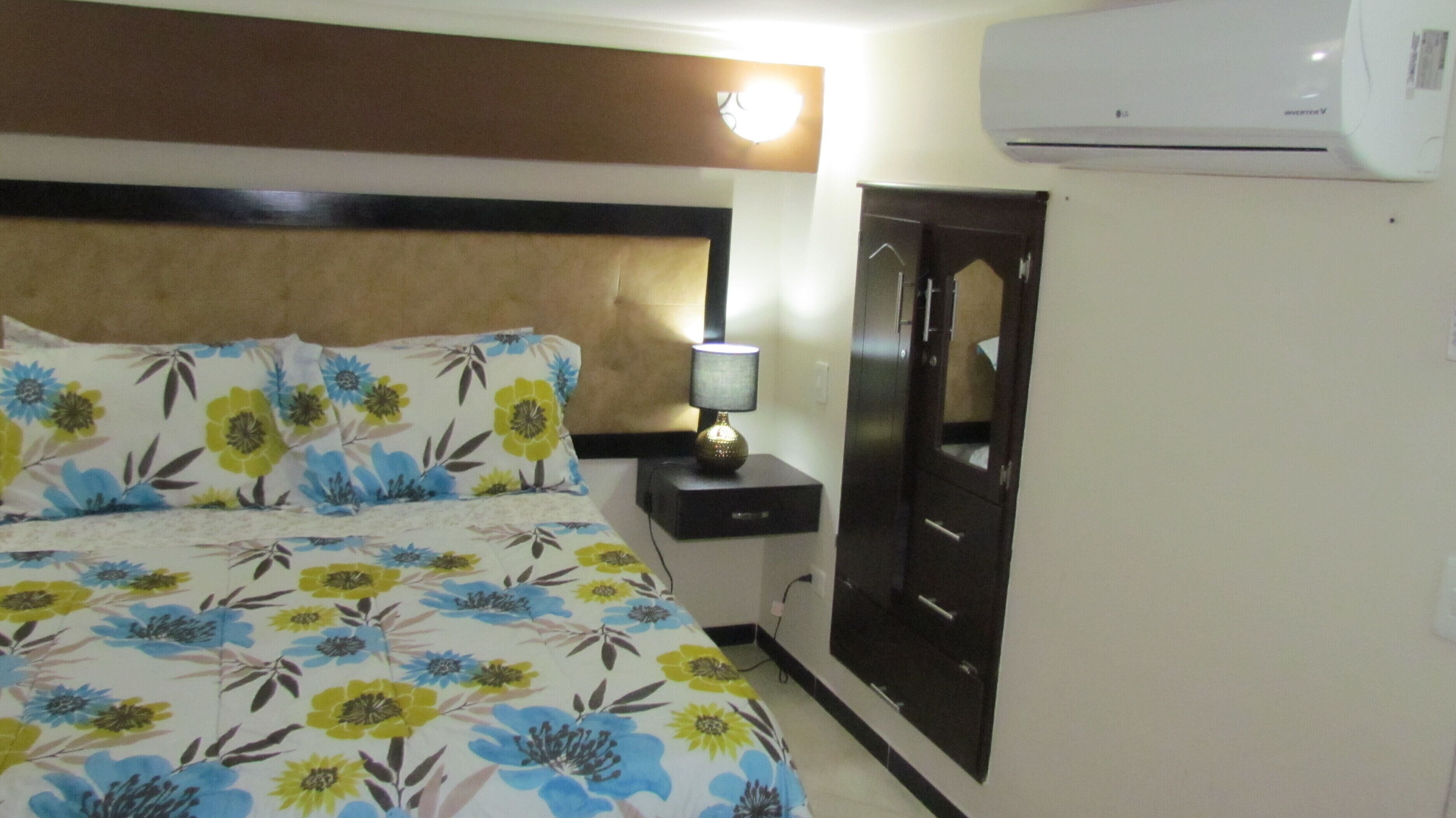 A hotel room with a bed, a nightstand, a lamp, a wardrobe, and an air conditioning unit.