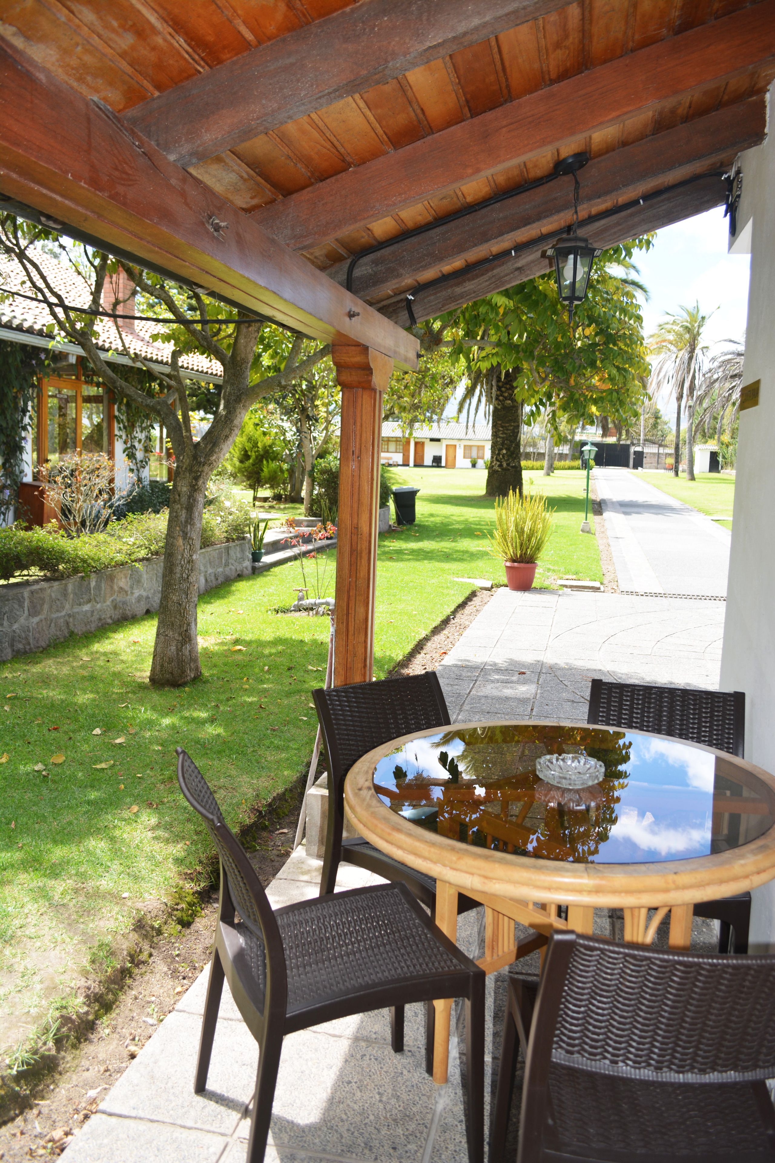 An outdoor patio with a round table and four chairs, surrounded by a stone wall and a grassy area with trees.