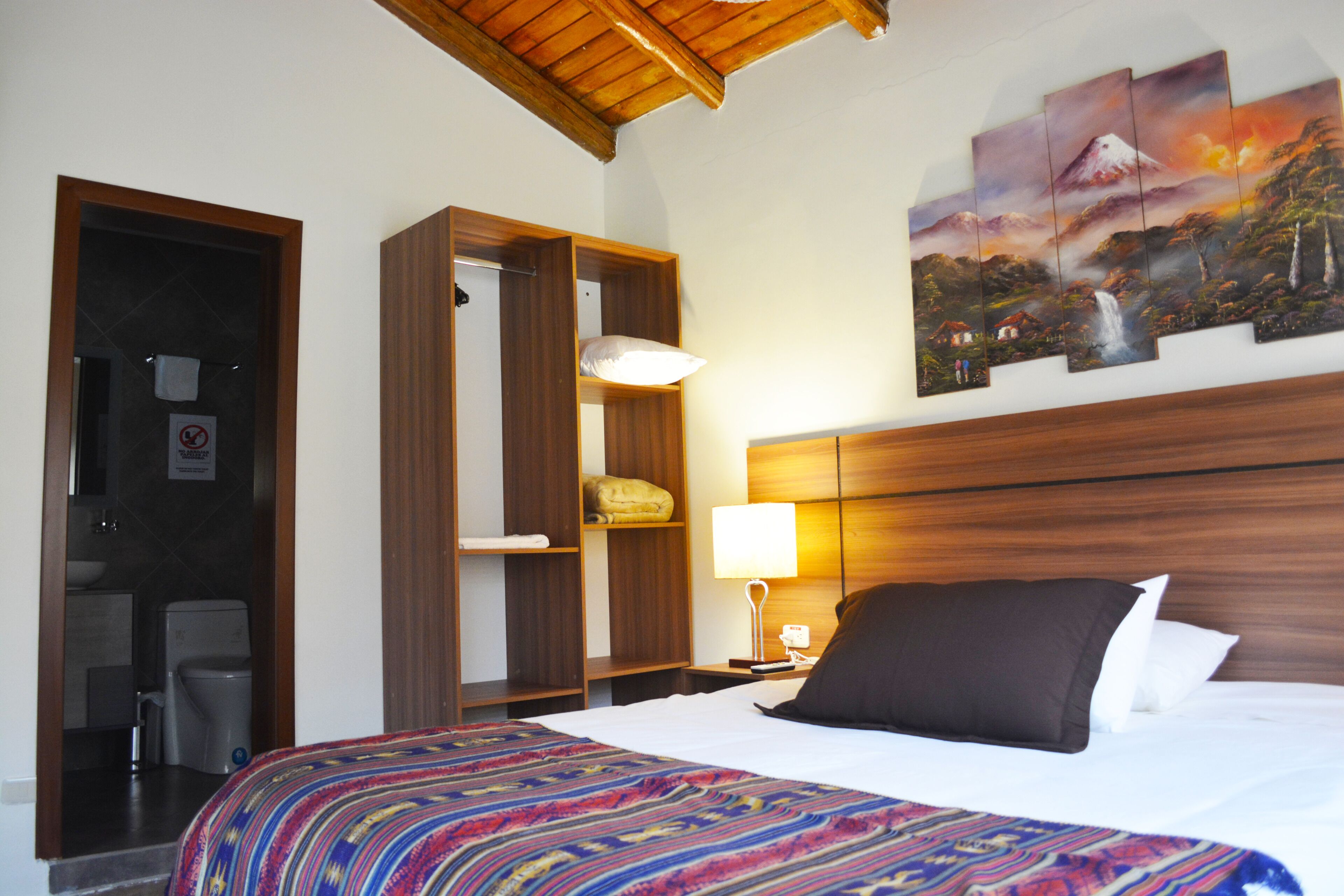 Une chambre d’hôtel comprenant un lit, une table de chevet avec une lampe, une tête de lit en bois, un tableau représentant un paysage de montagne et une armoire.