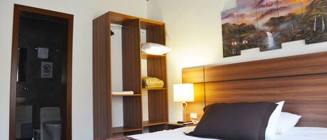 Une chambre d’hôtel comprenant un lit, une table de chevet avec une lampe, une tête de lit en bois, un tableau représentant un paysage de montagne et une armoire.