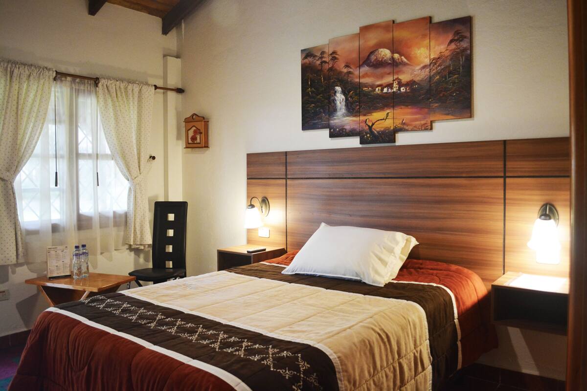 A hotel room with a large bed, two bedside lamps, a wooden headboard, a window with curtains, and a wall-mounted painting.