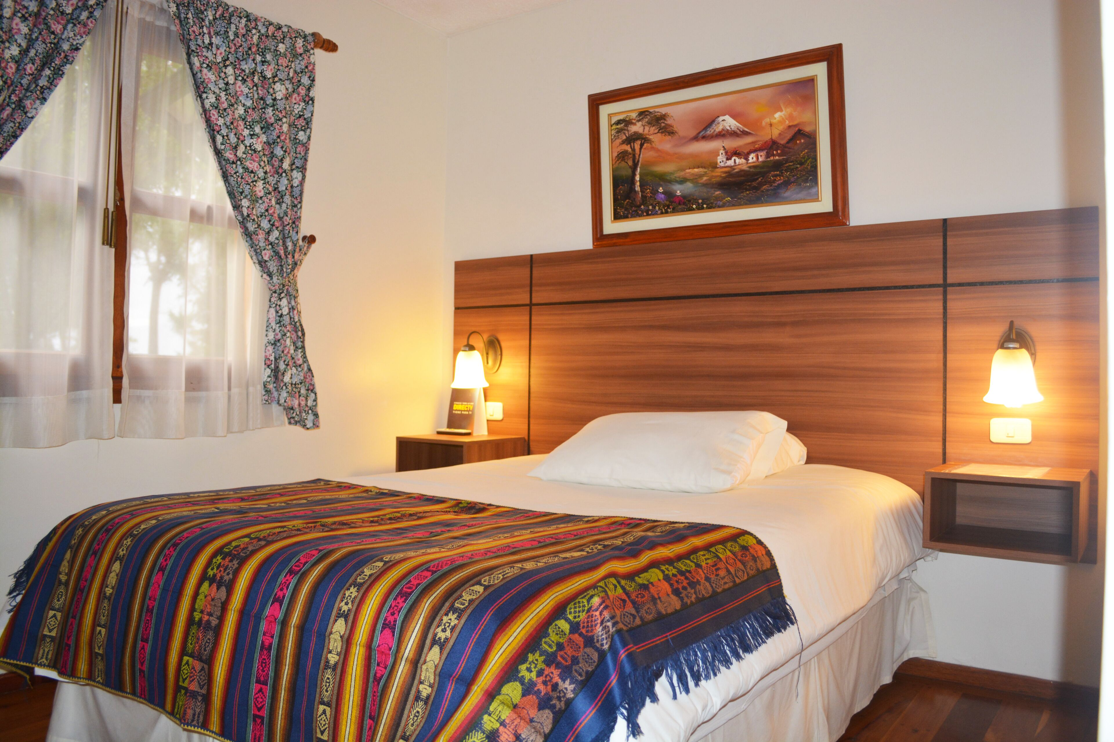 A hotel room with a wooden headboard, a colorful bedspread, two bedside lamps, and a framed picture on the wall.