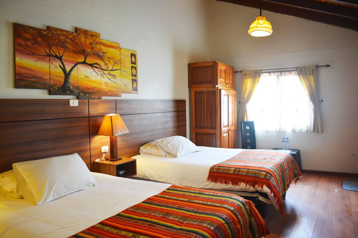 A hotel room with two beds, wooden headboard, a nightstand with a lamp, a wardrobe, and a window with curtains.
