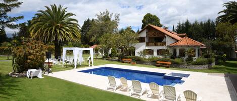 A swimming pool with lounge chairs, a white gazebo, and a house with a tiled roof.
