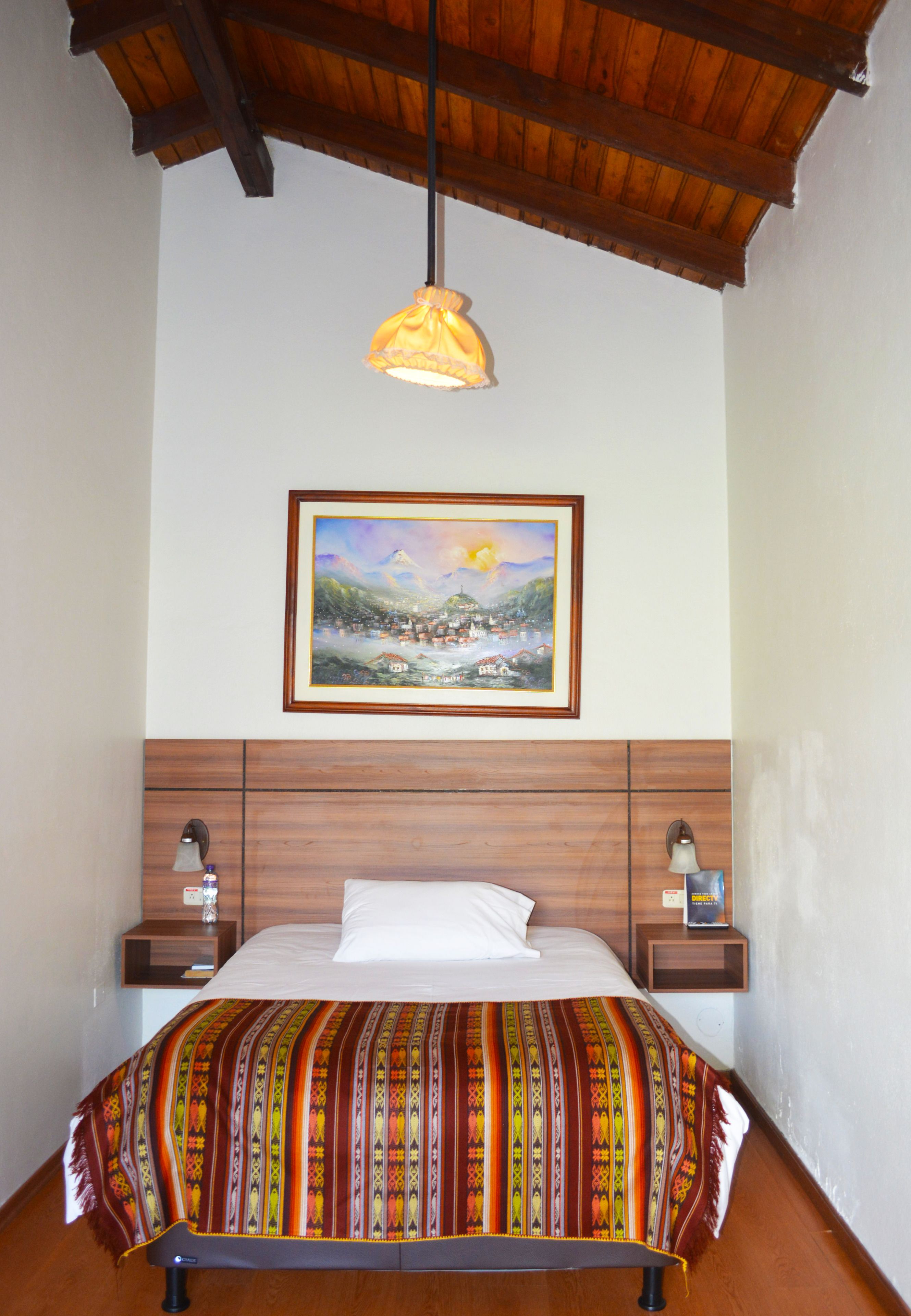 A neatly made bed with a colorful blanket, a framed painting above it, and a wooden ceiling.