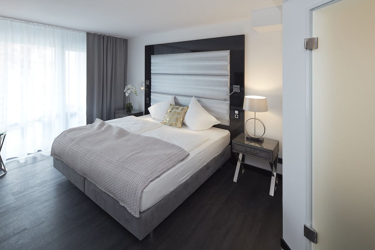 A modern hotel room with a large bed, a bedside table with a lamp, and a view of the outdoors.