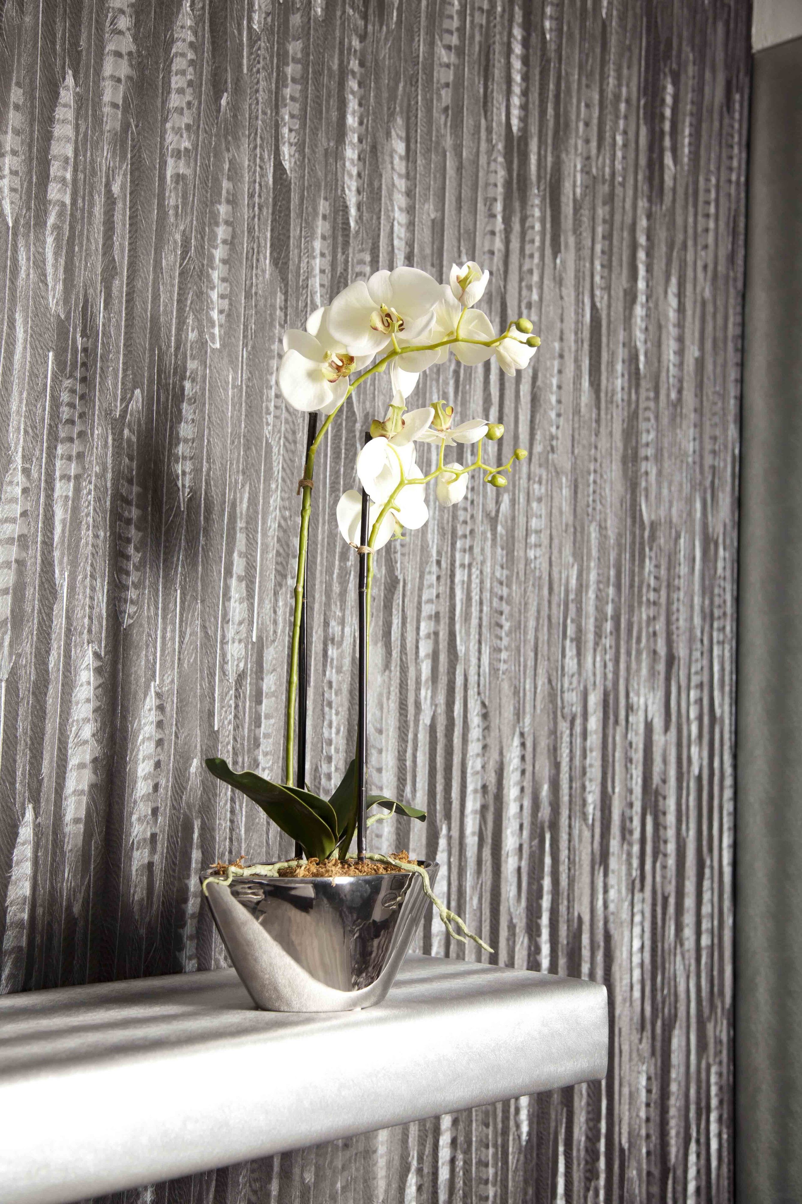 Una maceta de plata con orquídeas sobre un estante blanco frente a una pared gris con textura.