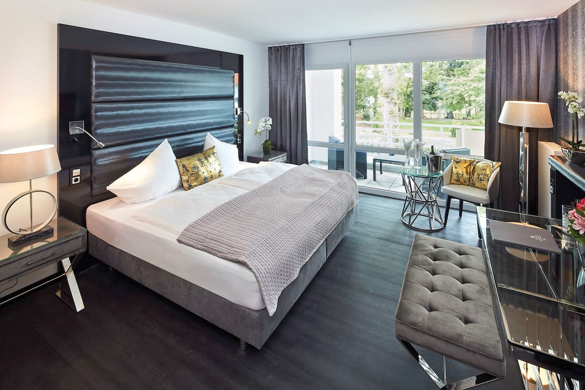 A modern hotel room with a large bed, a glass dining table, and a view of the outdoors.