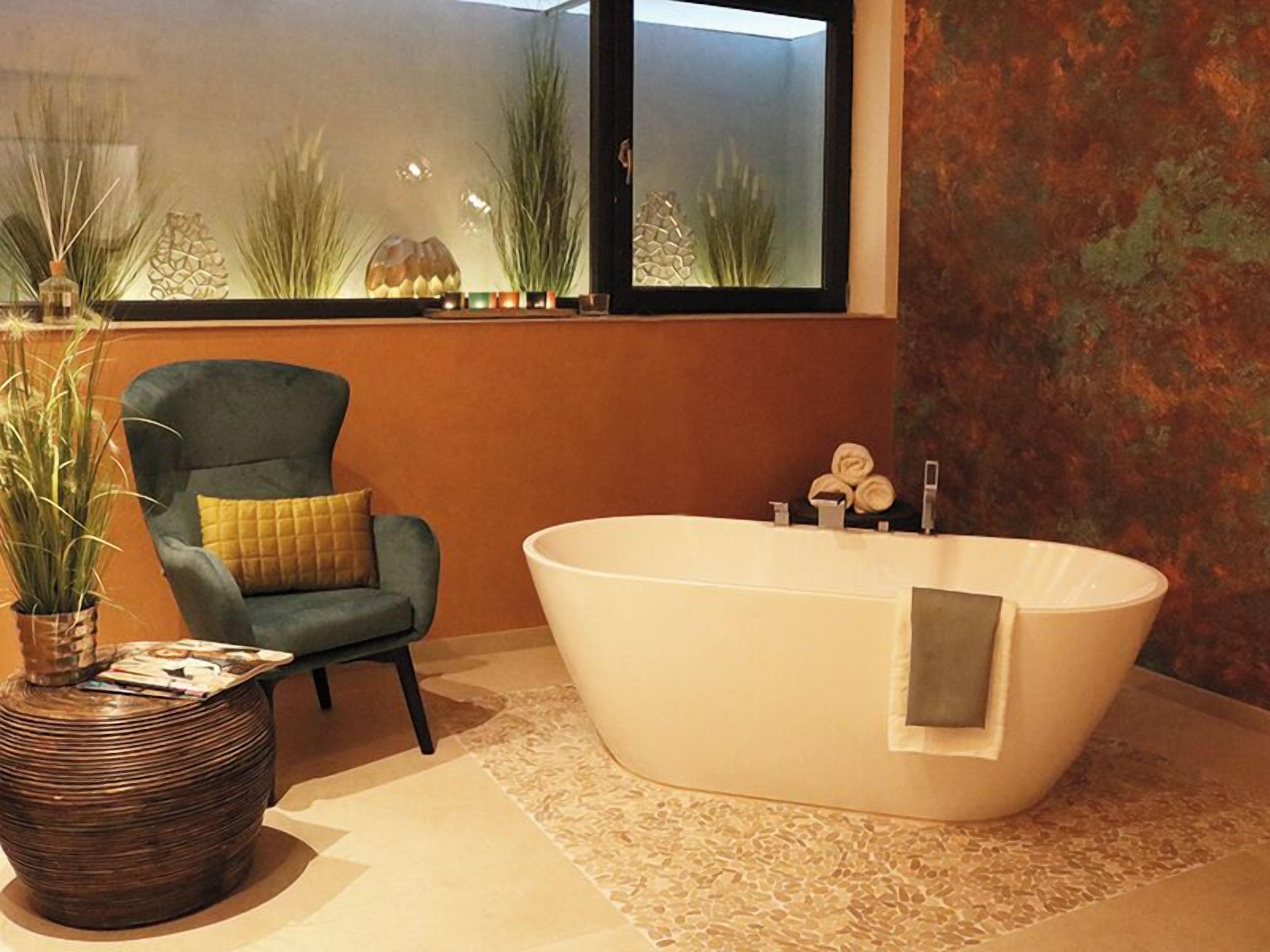 Un baño moderno con una bañera independiente, un sillón verde y un amplio ventanal con plantas.