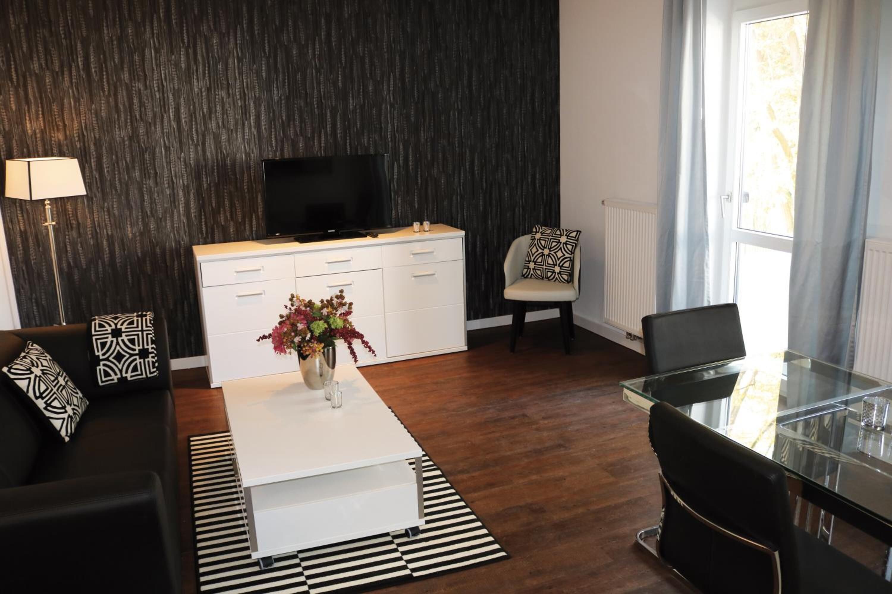 Una sala de estar moderna con un sofá blanco, una mesa de centro de vidrio, un televisor negro sobre un mueble blanco, y un comedor con una mesa y sillas de vidrio.