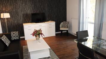 Una sala de estar moderna con un sofá blanco, una mesa de centro de vidrio, un televisor negro sobre un mueble blanco, y un comedor con una mesa y sillas de vidrio.