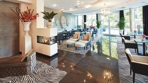 A spacious lobby with a fireplace, seating area, and large windows offering a view of the outdoors. - Kurhaus Design Boutique Hotel (Erwitte)