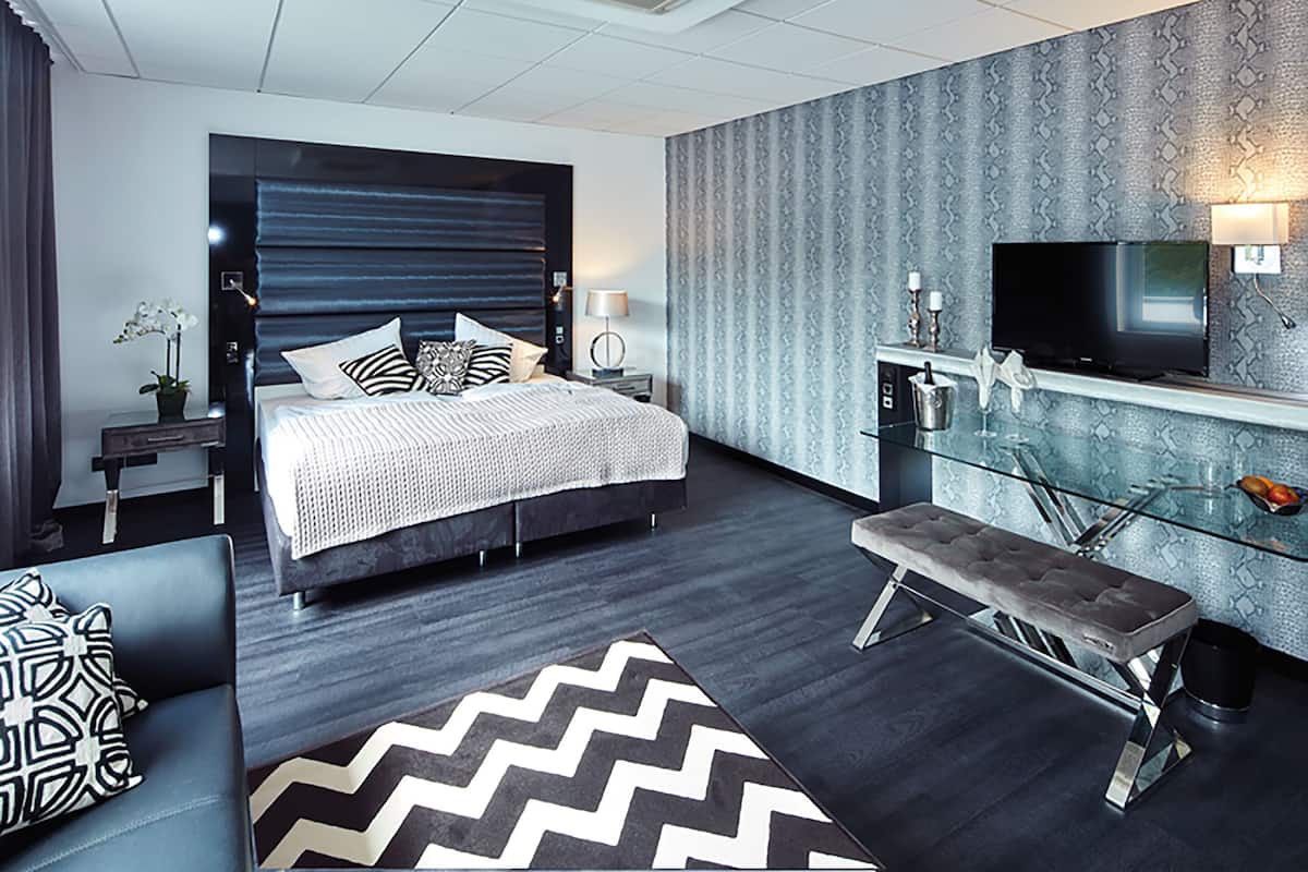 A modern hotel room with a large bed, a TV, a desk, and a bench.