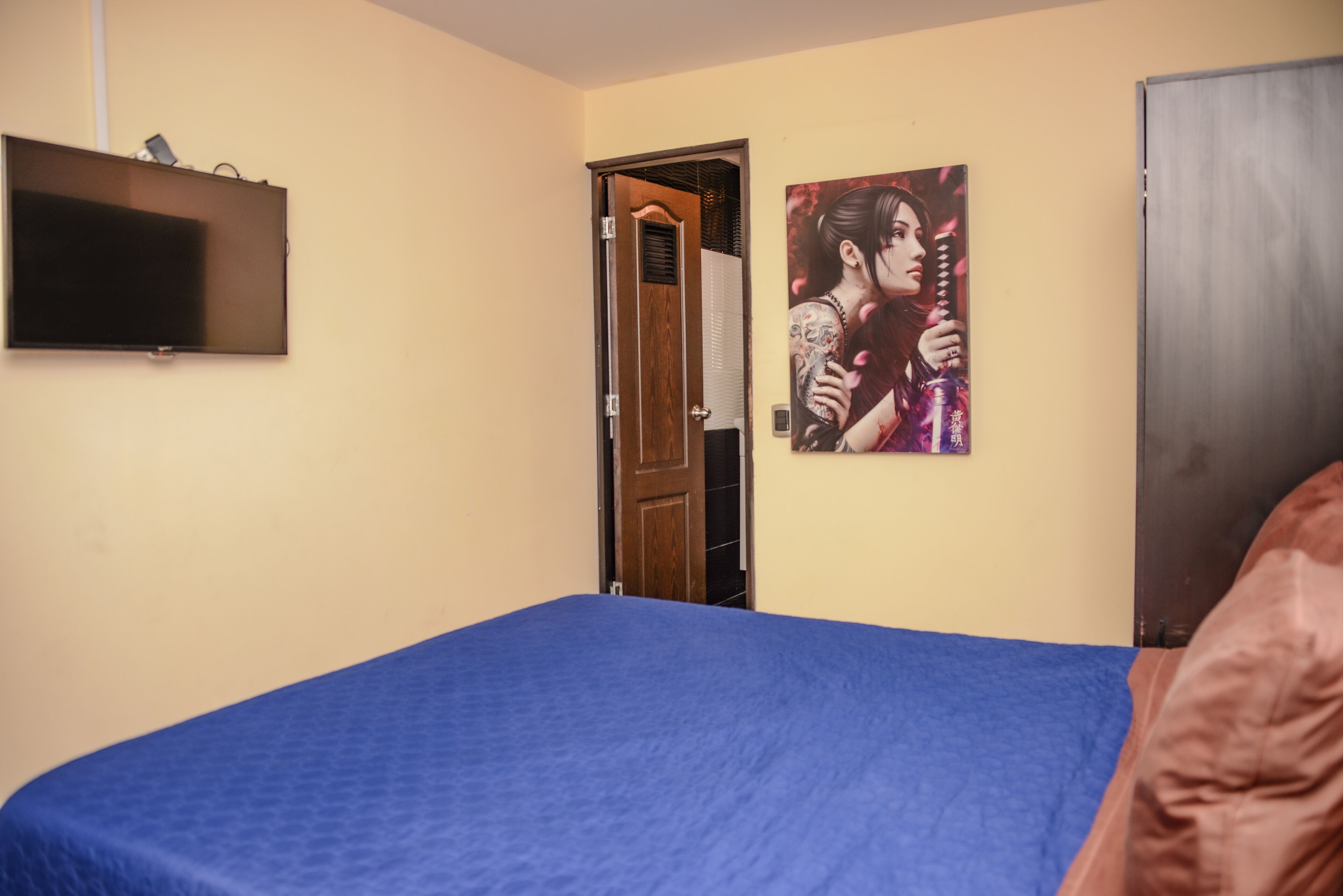 Ein Schlafzimmer mit einem Bett, einem an der Wand befestigten Fernseher und einer Tür zu einem weiteren Raum.