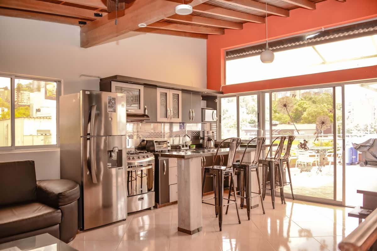 A modern kitchen with stainless steel appliances, a central island with bar stools, and large windows offering a view of the outdoors.