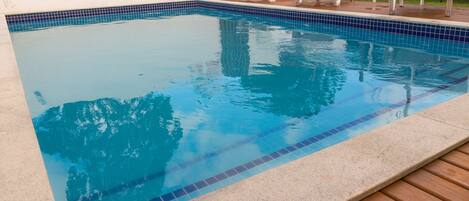 Piscina rectangular con azulejos azules, rodeada por una terraza de madera y sillas reclinables blancas.