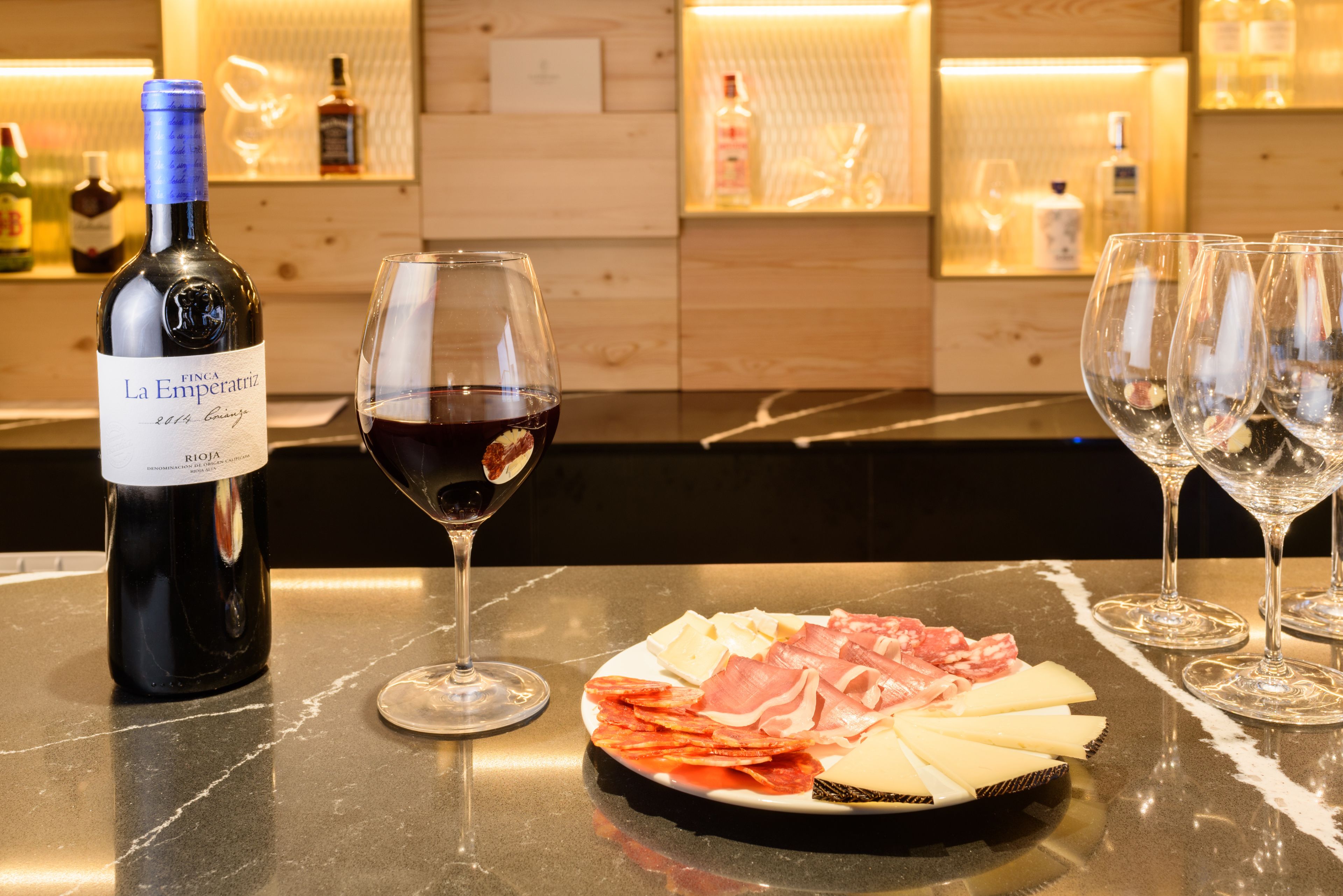 Zona de bar con una botella de vino, una copa de vino tinto y una bandeja de quesos y embutidos.