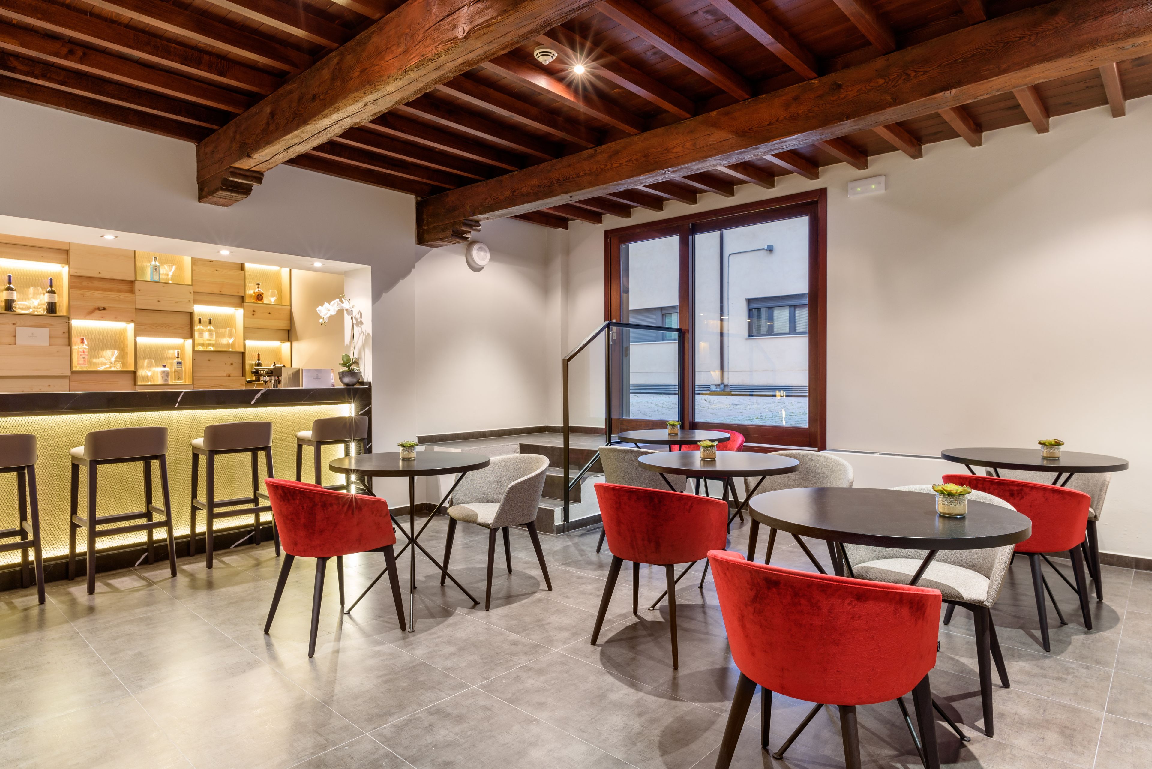 A modern bar area with wooden beams, a bar counter with stools, and round tables with red chairs.