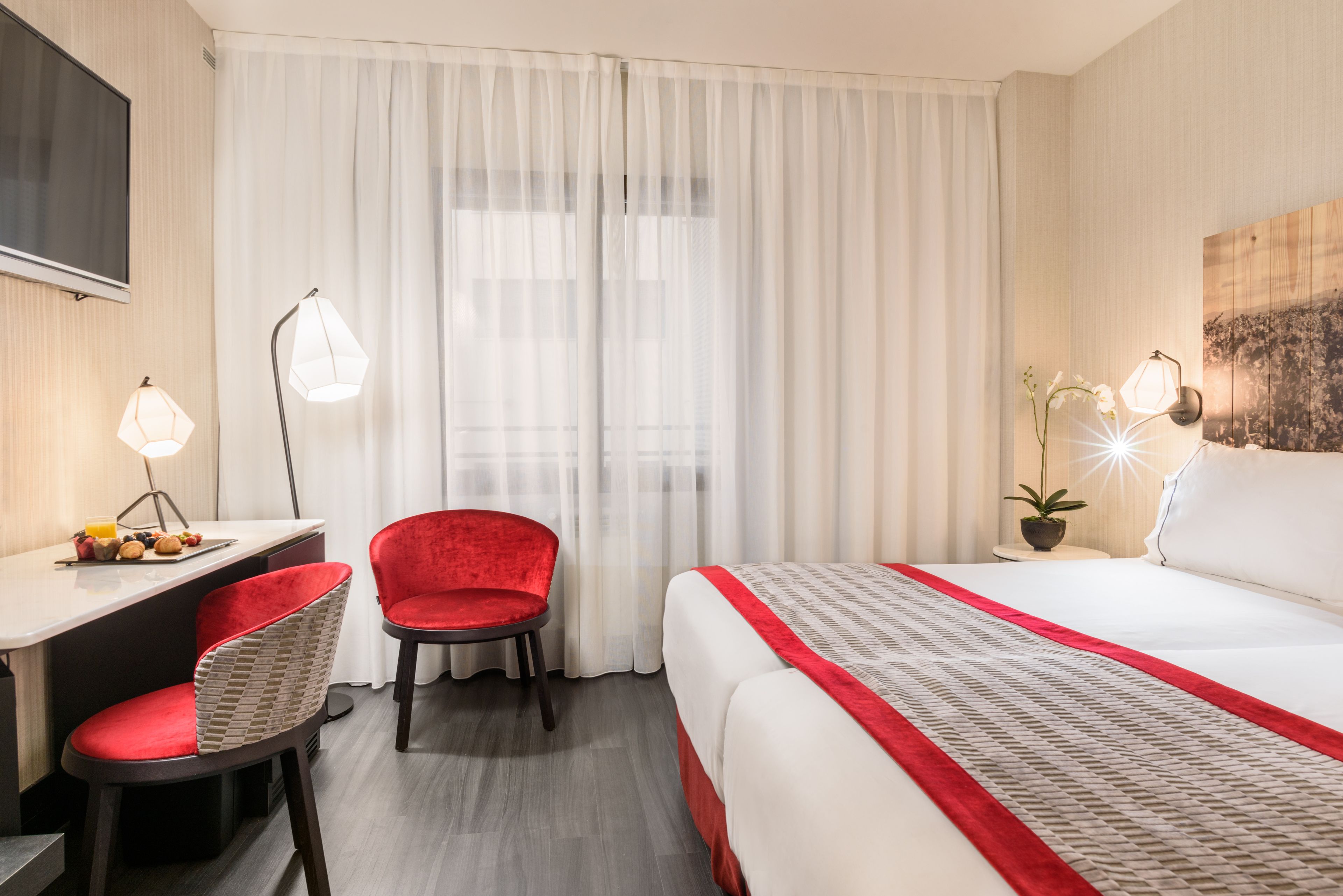 A hotel room with a bed, a desk, a chair, a TV, and a window with curtains.