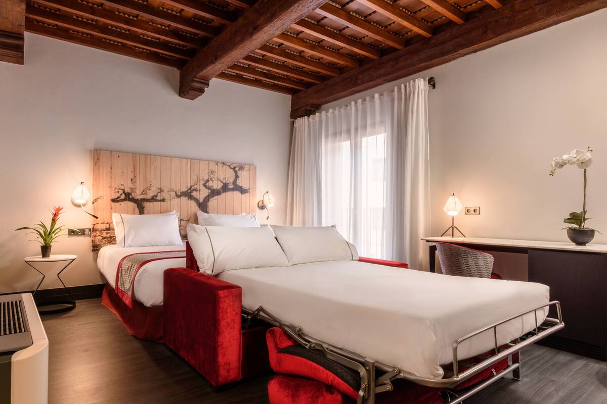 Une chambre d’hôtel comprenant un lit, un bureau, une chaise, une plante en pot et une œuvre d’art accrochée au mur.