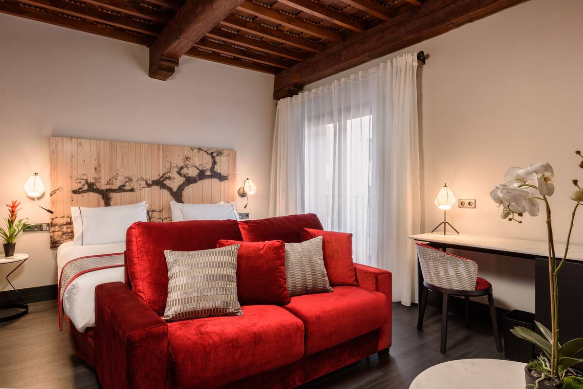 Une pièce chaleureuse avec un canapé rouge, un lit recouvert de draps blancs, une œuvre d’art murale en bois, un bureau avec une chaise et une plante orchidée en pot.