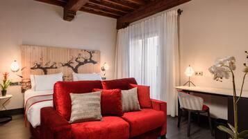 A cozy room with a red sofa, a bed with white linens, a wooden wall art piece, a desk with a chair, and a potted orchid plant.
