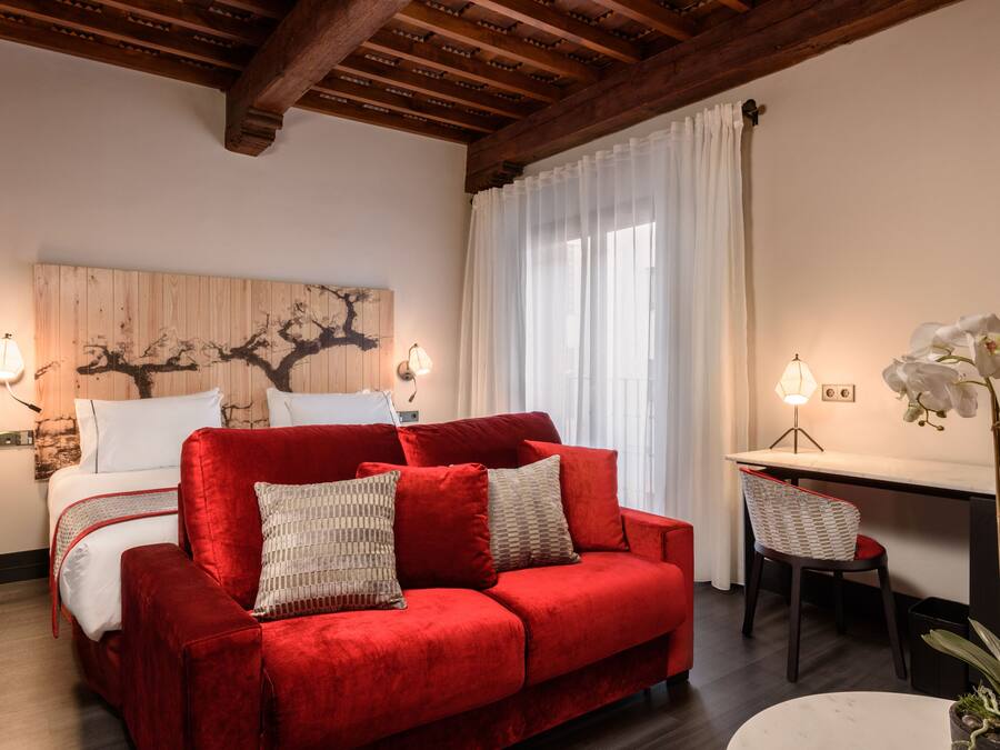 A cozy room with a red sofa, a bed with white linens, a wooden wall art piece, a desk with a chair, and a potted orchid plant.