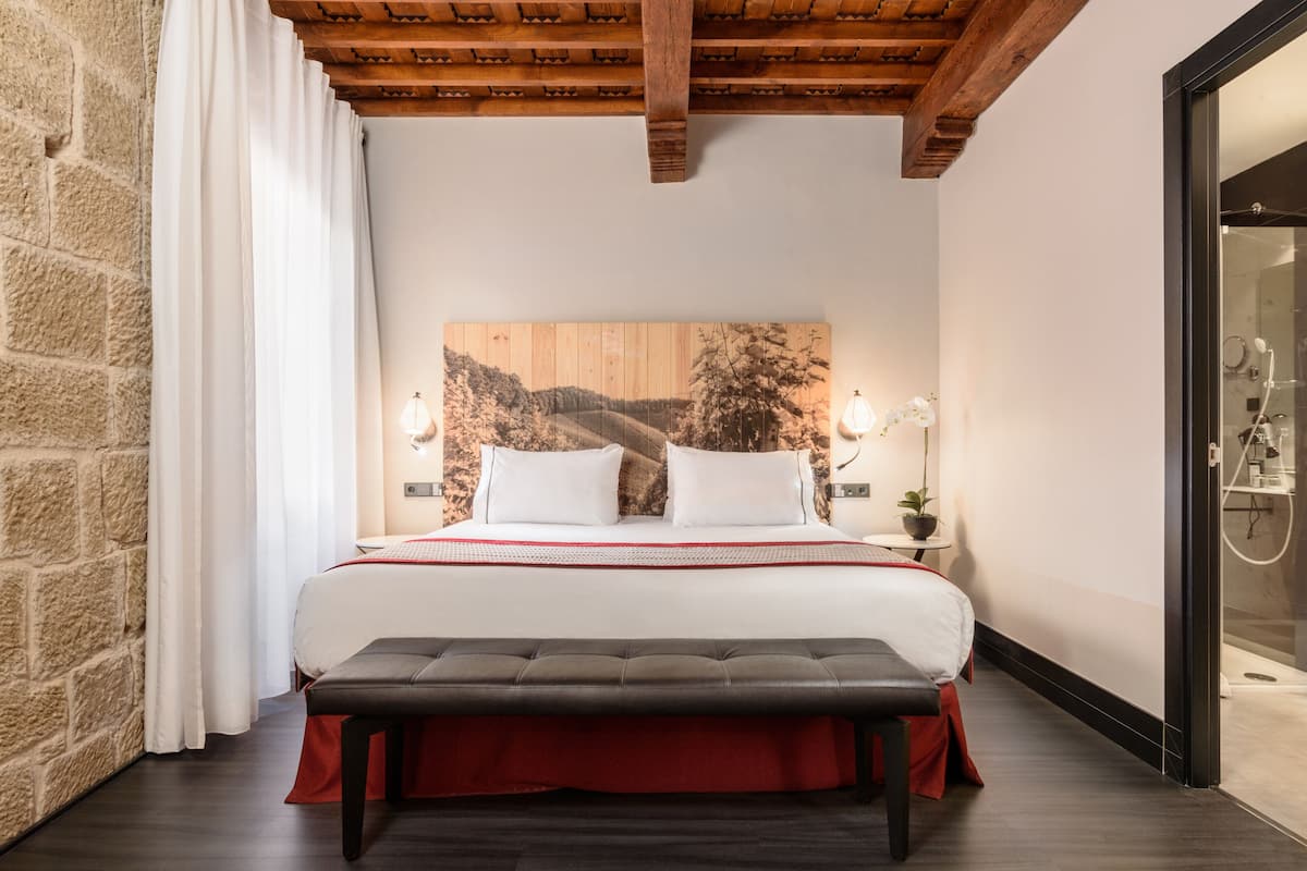 Une chambre avec un grand lit, une tête de lit en bois, un banc, une table de chevet avec une lampe et une vue sur la salle de bain.