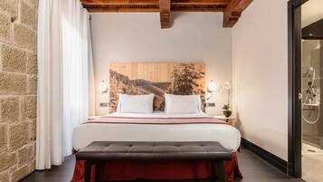 Un dormitorio con una cama grande, cabecera de madera, un banco, una mesita de noche con lámpara y vistas al baño.