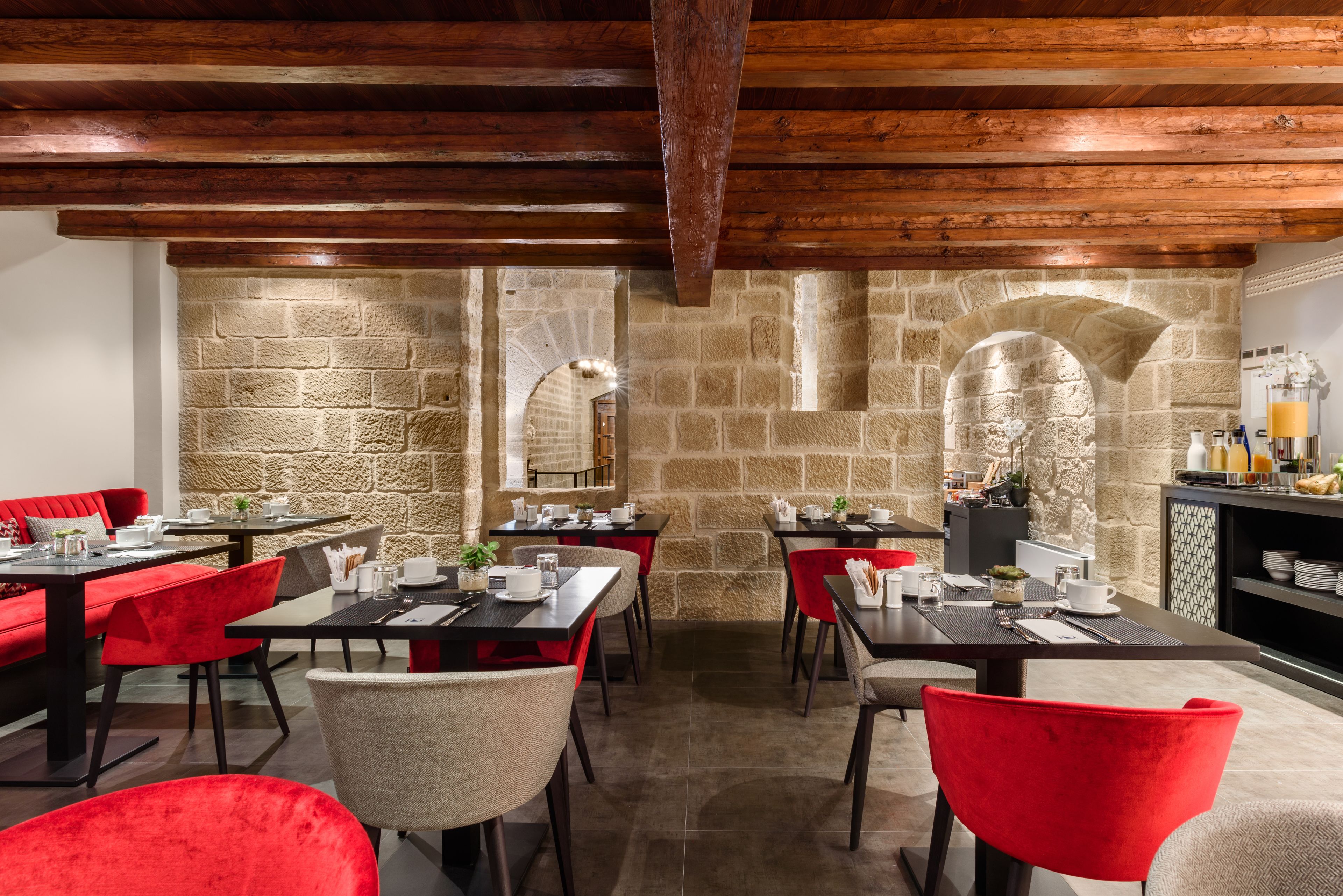 A restaurant with stone walls, wooden beams, and red chairs. Tables are set with plates, glasses, and cutlery.