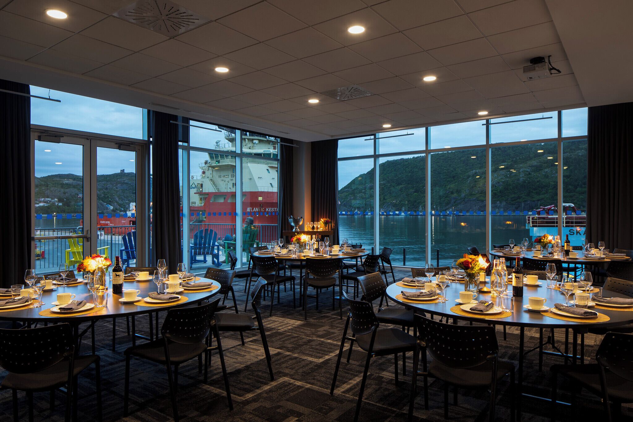 Un ristorante con diversi tavoli rotondi apparecchiati per la cena, ampie finestre che offrono vista su una nave e delle colline, e un design moderno del soffitto.