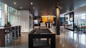 A modern indoor pool table area with bar seating and a bar counter, surrounded by large windows and a reception area. - Alt Hotel St. John's (St. John's)