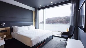 Een moderne hotelkamer met een groot bed, een bureau en een stoel. Het heeft een groot raam met uitzicht op een wateroppervlak en heuvels.