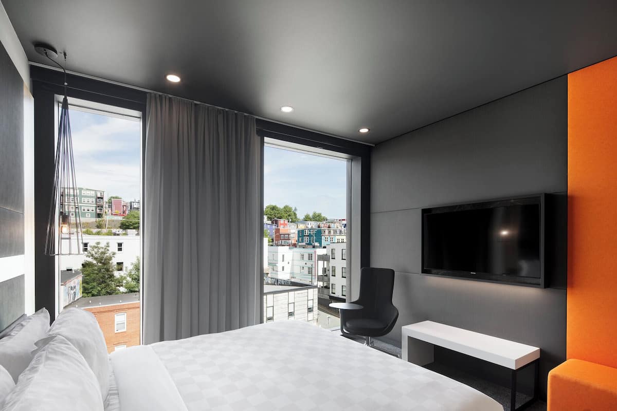 A modern hotel room with a large bed, a TV mounted on the wall, a bench, and a view of the cityscape through large windows.