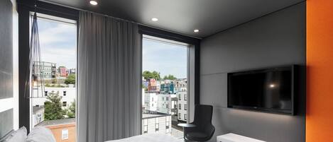 A modern hotel room with a large bed, a TV mounted on the wall, a bench, and a view of the cityscape through large windows.